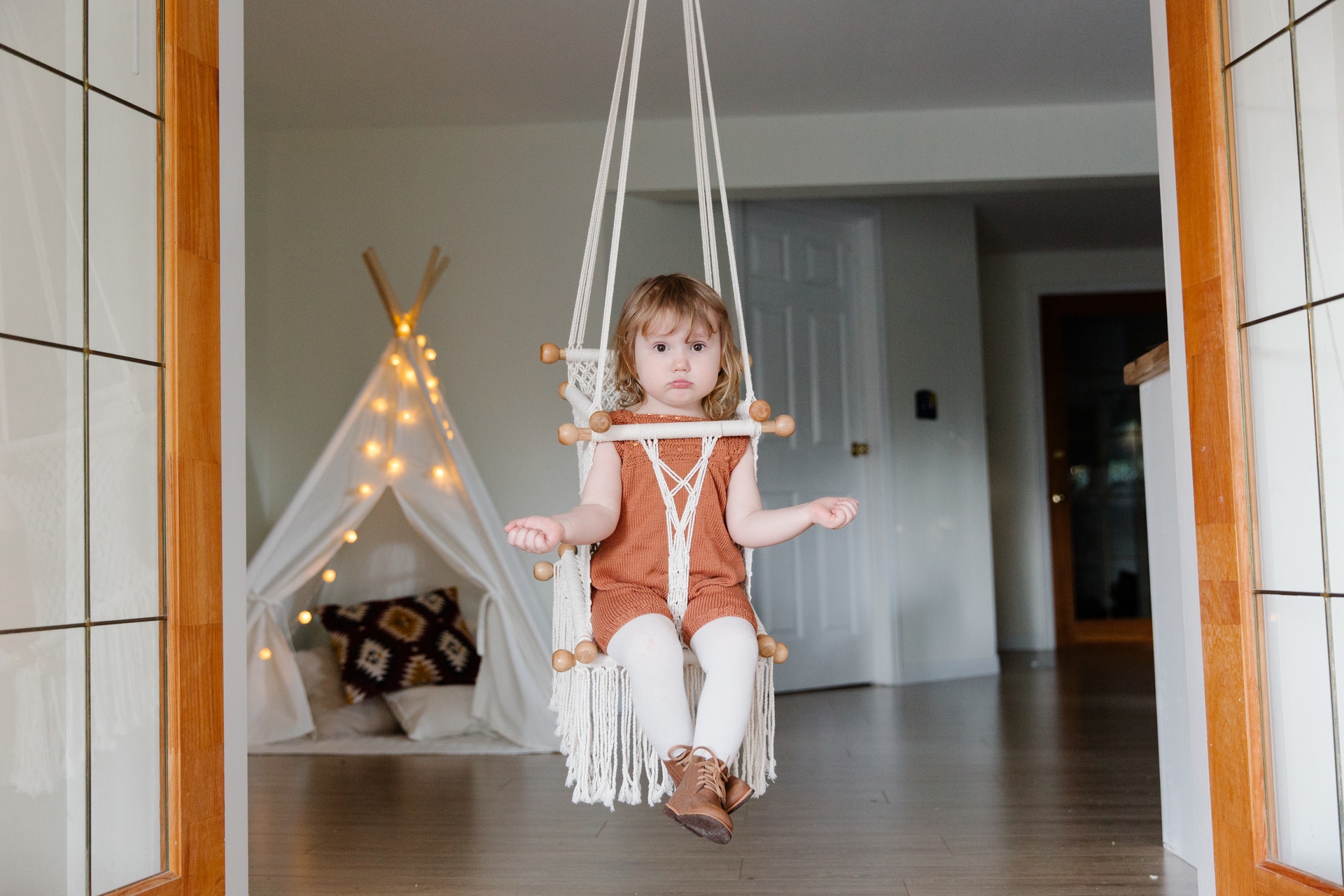Right or wrong for you - small female child sitting in a rope swing in a doorway shrugging with a tent and fairy lights behind her